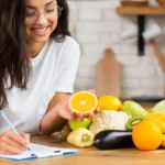 mulher de óculos feliz anotando algo com uma fruta na mão e ingredientes frutas e vegetais no balcão para hipercalórico caseiro e ganho de massa