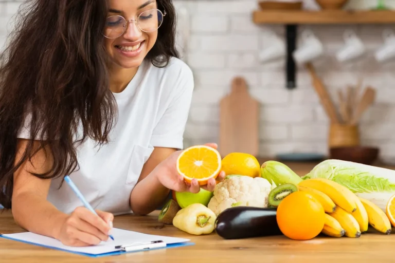 mulher de óculos feliz anotando algo com uma fruta na mão e ingredientes frutas e vegetais no balcão para hipercalórico caseiro e ganho de massa